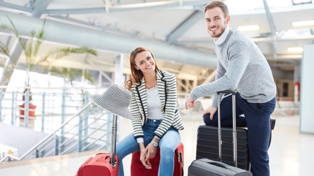 A imagem mostra um casal feliz e sorridente com suas malas no saguão do aeroporto próximo à porta