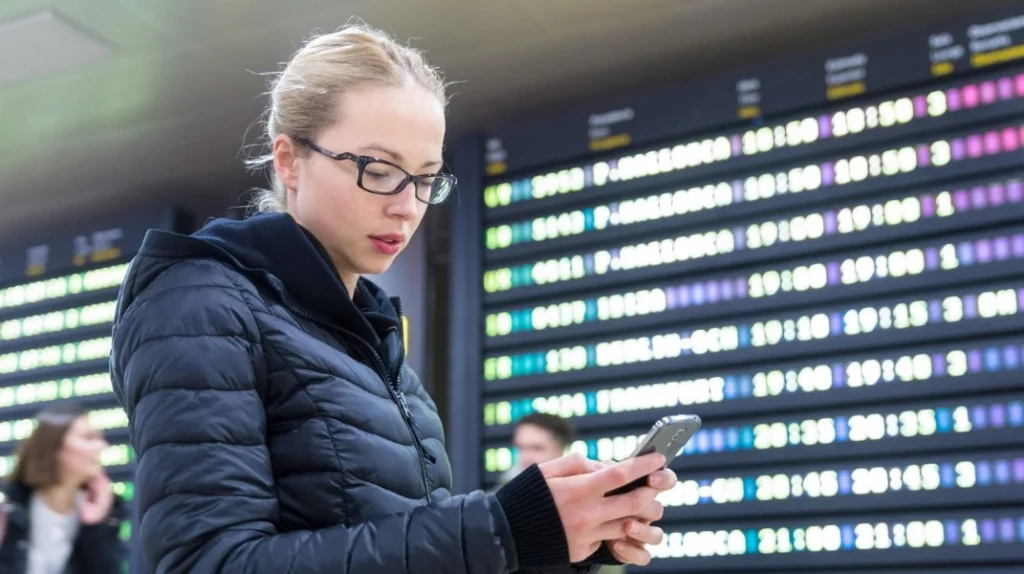 A imagem mostra uma passageira no saguão do aeroporto verificando dados do voo no aplicativo de celular enquanto aguarda seu avião