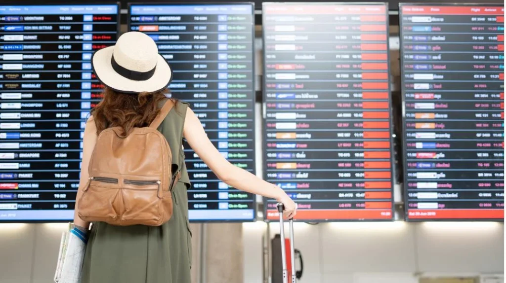 A imagem mostra uma passageira acompanhando o painel de partidas e chegadas no saguão do aeroporto enquanto aguarda seu avião