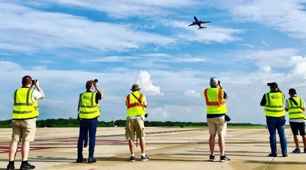 A imagem mostra spotters credenciados acompanhando e fotografando a decolagem de grandes aviões próximo de um aeroporto. Estão praticando plane spotting como hobby